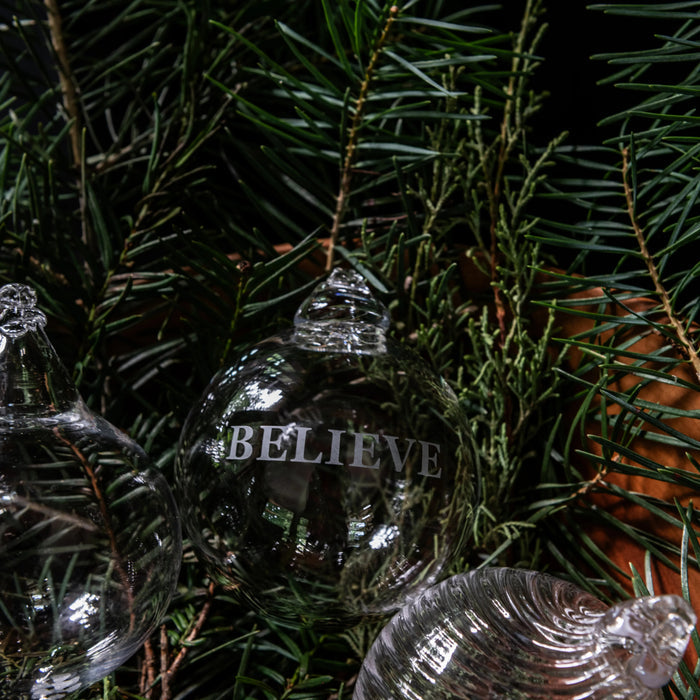 Black glossy round ornament engraved with "BELIEVE" text hanging among evergreen branches.