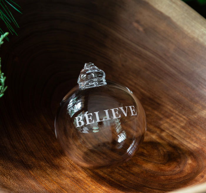 Clear glass round ornament with "BELIEVE" engraved text and metallic cap, displayed on wood surface.