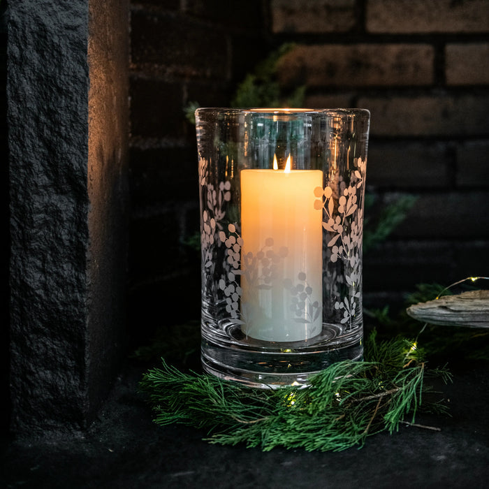 Clear glass hurricane with engraved berry and leaf pattern, containing lit cream-colored pillar candle.