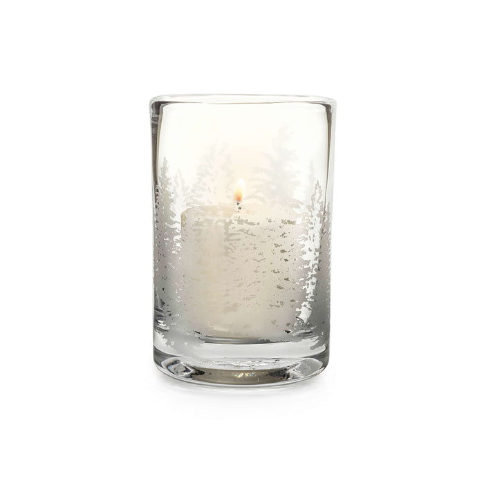 Clear glass hurricane candle with evergreen tree engraving and lit cream-colored candle inside.