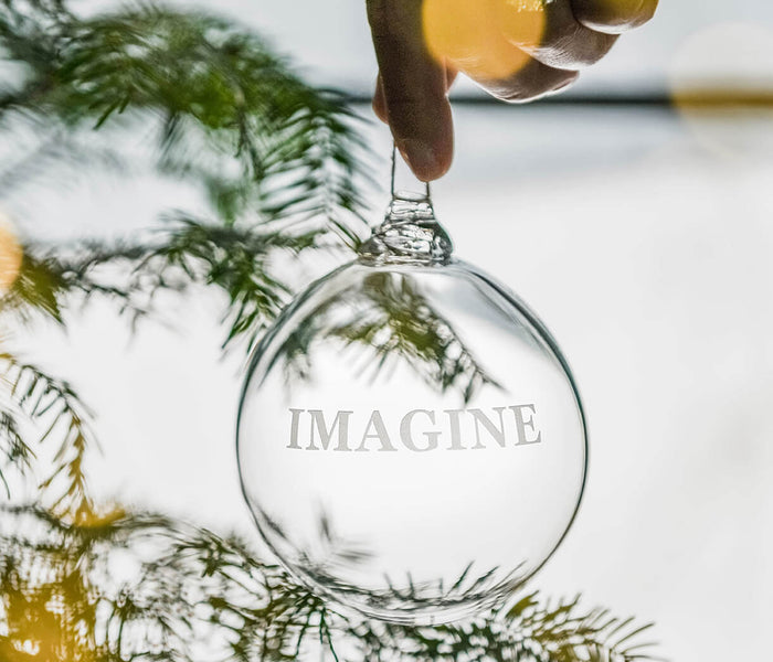 Clear glass round ornament engraved with "IMAGINE" text, hanging from a gold-toned cap on evergreen branch.