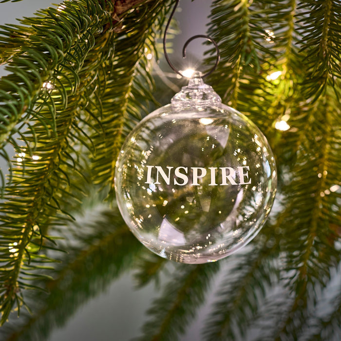 Clear glass round ornament engraved with "Inspire" text, hanging on evergreen branch with metal hook.