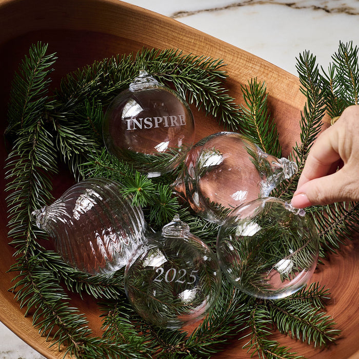Round glass ornaments engraved with "Inspire" and "2025" nestled in evergreen branches in a wooden bowl.