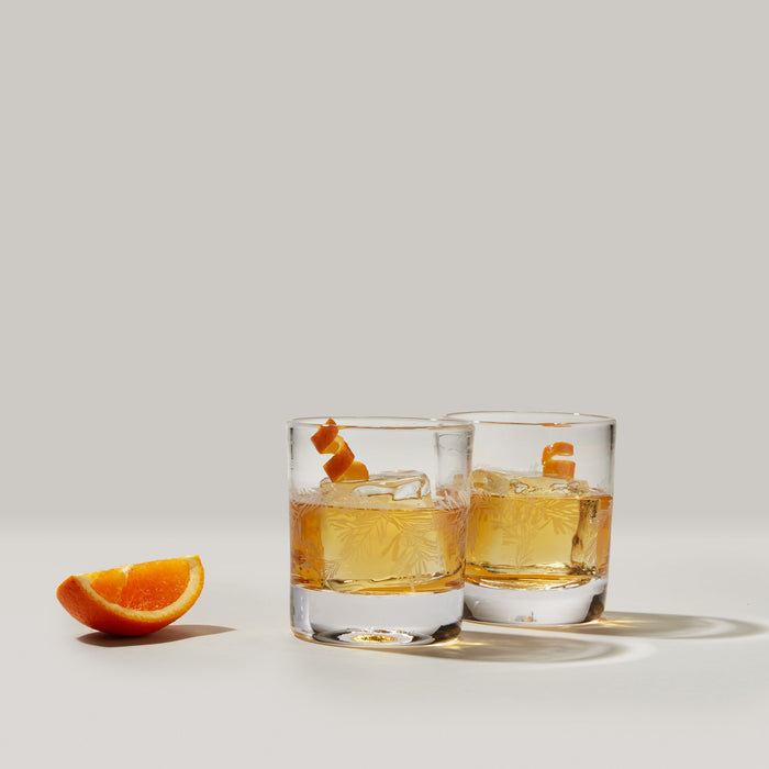 Two clear double old-fashioned glasses filled with amber liquid and ice, with orange slices and peel garnish.