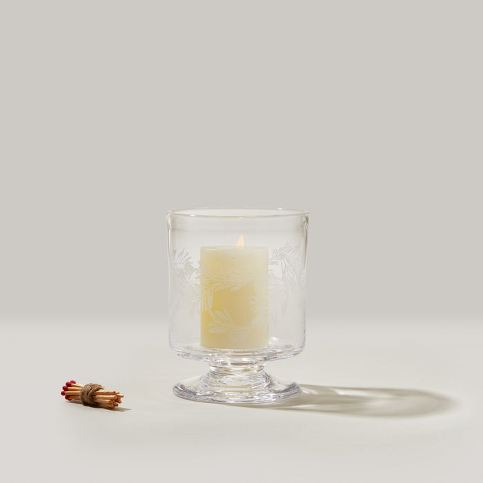 Clear pedestal hurricane with engraved botanical design holding a lit cream candle next to bundled cinnamon sticks.