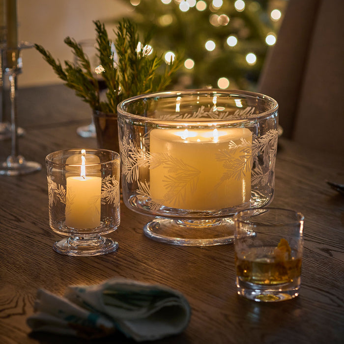 Clear glass hurricane candleholders with engraved pine branch design, small and large sizes, each containing a lit cream cand