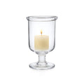 Clear glass hurricane vase with pedestal base holding a lit cream-colored pillar candle.
