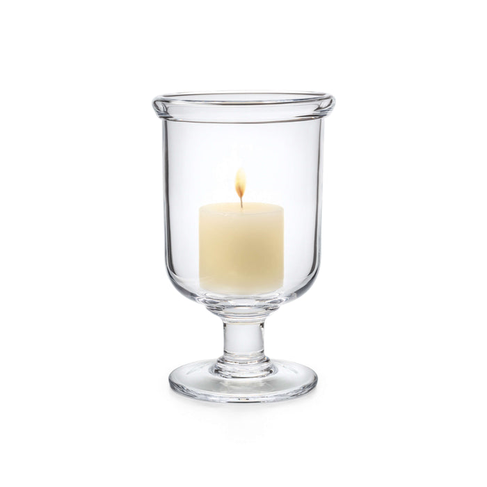 Clear glass hurricane vase with pedestal base holding a lit cream-colored pillar candle.