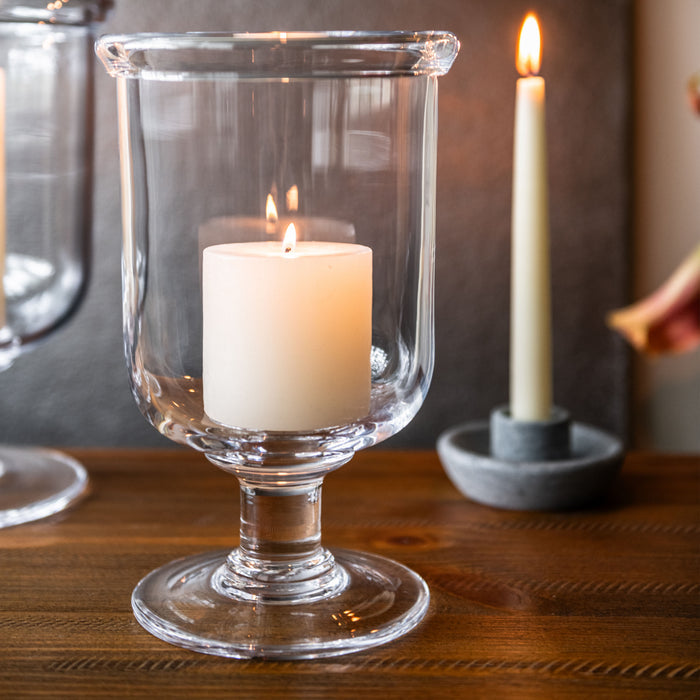 Clear glass hurricane candle holder with pedestal base containing white pillar candle.