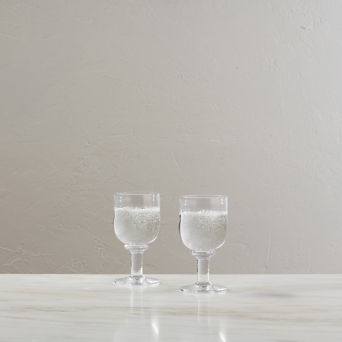 Two clear water glasses with stems, each filled with sparkling water, displayed on a white surface against a neutral backgrou