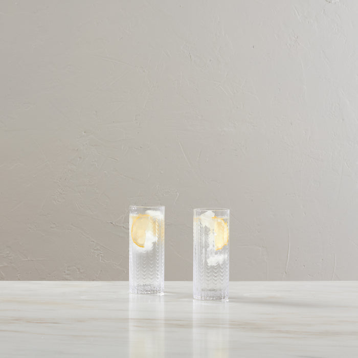 Two tall Collins glasses with textured pattern, each containing ice and lemon slices in clear liquid.