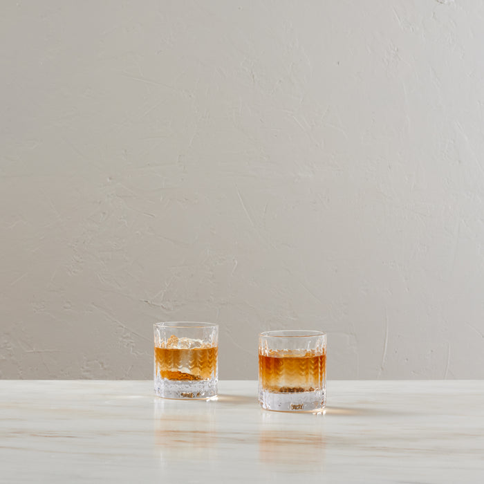 Two clear lowball glasses filled with amber liquid and ice on a white surface.