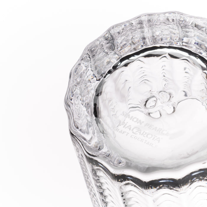 A close-up of a clear textured glass tumbler, showing the inside base engraved with 'Simon Pearce Via Carota Craft Cocktails' on a white background.
