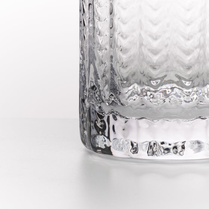 A close-up of a clear textured glass tumbler with a wavy, hammered pattern and a thick ridged base, set against a clean white background.