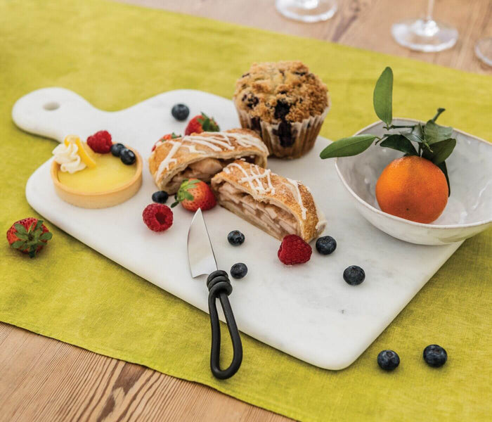 Cheese knife with black handle on white marble serving board with pastries and berries.