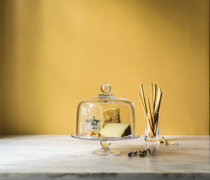 Glass cake plate with dome cover on pedestal base, displayed with sliced cake and gold serving utensils.