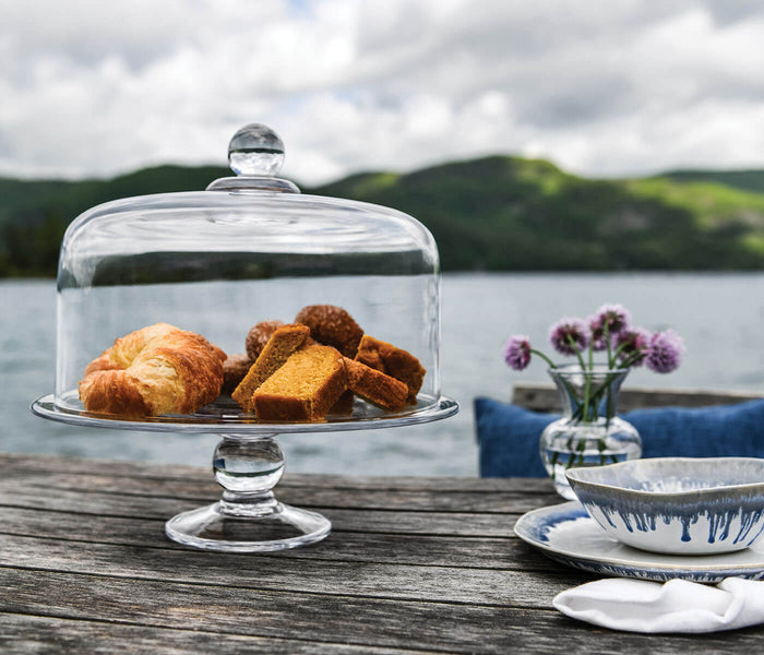 Glass cake plate with dome lid on pedestal base, filled with baked goods, displayed outdoors by water.