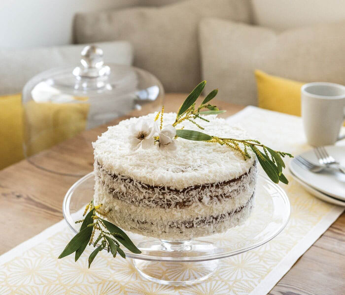 Clear glass cakeplate with pedestal base holding a layered cake topped with greenery and white flowers.