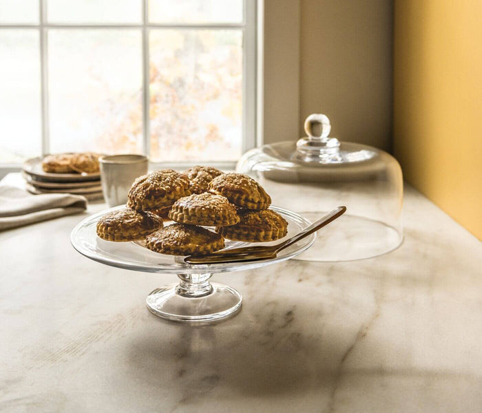Clear glass cake plate with pedestal base holding baked goods on marble table.
