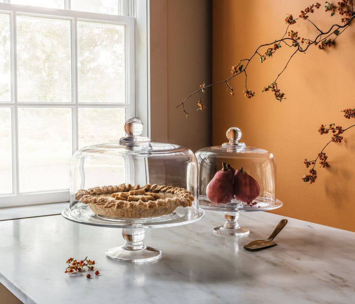 Clear glass cake plate with domed lid on pedestal base, displayed with pie and fruit.