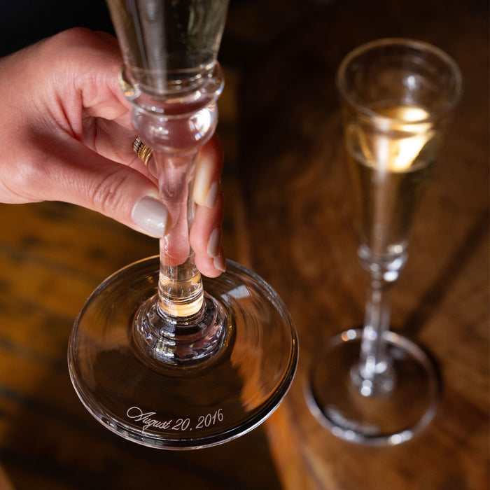Clear champagne flute with gold-trimmed base, engraved "August 20, 2016," held in hand with additional flutes visible.