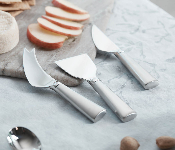 Three cheese knives with white handles and silver blades arranged on marble surface with sliced cheese and nuts.