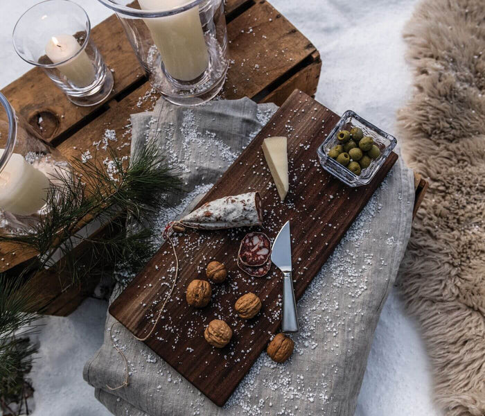 Cheese knife with metal blade and handle displayed on wooden board with cheese, walnuts, olives, and candles.