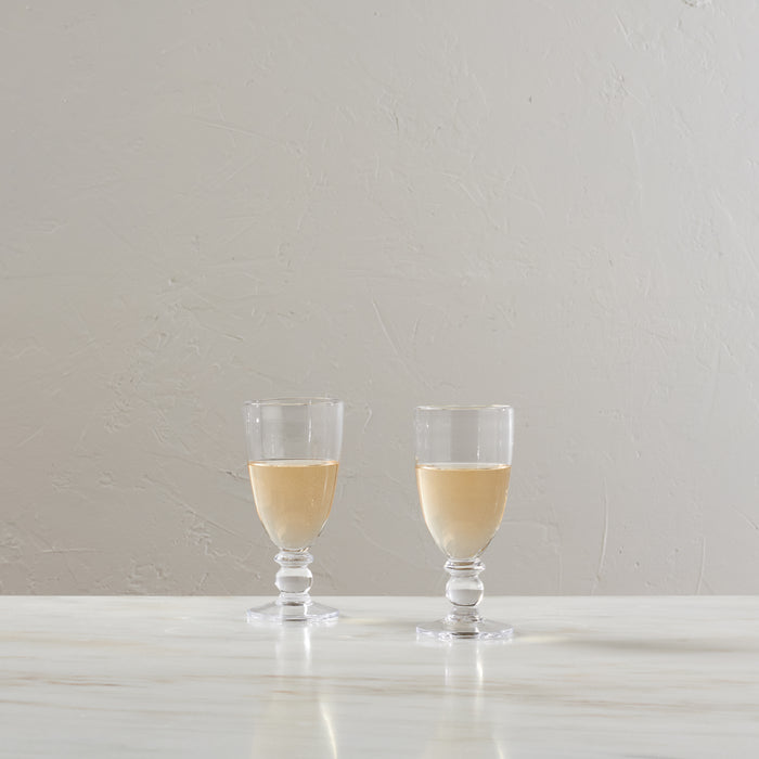 Two stemmed glasses filled with pale yellow wine on a white surface against a beige textured wall.