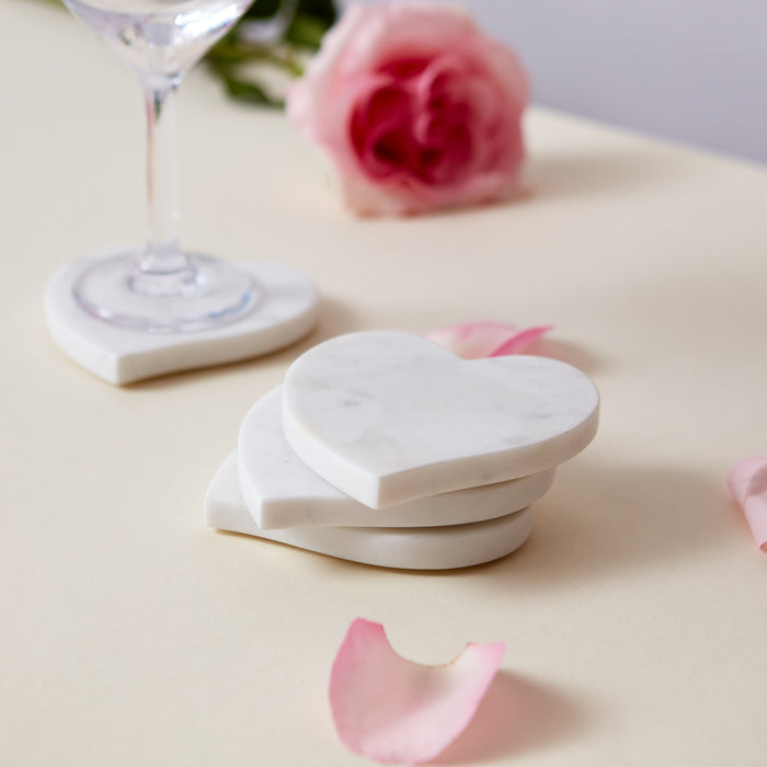 Three white marble heart-shaped coasters stacked together on a surface with decorative flowers and glassware.