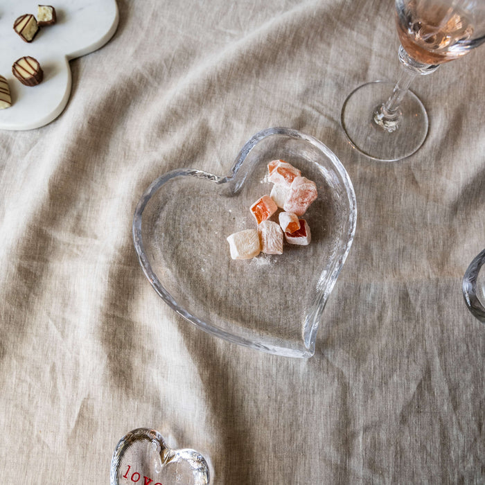 Clear glass heart-shaped tray holding pink and white candies on beige linen surface.
