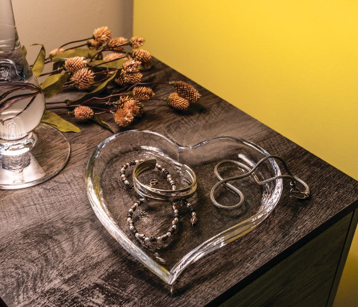 Clear glass heart-shaped tray holding silver bracelets and beaded jewelry on dark wooden surface.