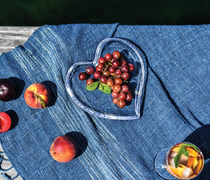 Clear heart-shaped tray filled with red grapes, placed on blue fabric with fresh fruit nearby.