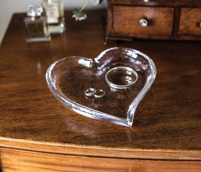 Clear glass heart-shaped tray with small ring holder compartment on wooden dresser.