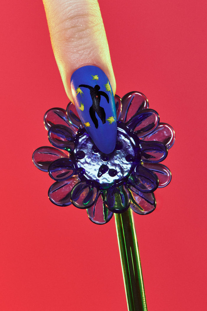 Henri Matisse Icarus almond press-on nail: blue with Icarus silhouette & yellow stars. Held by finger over purple flower.