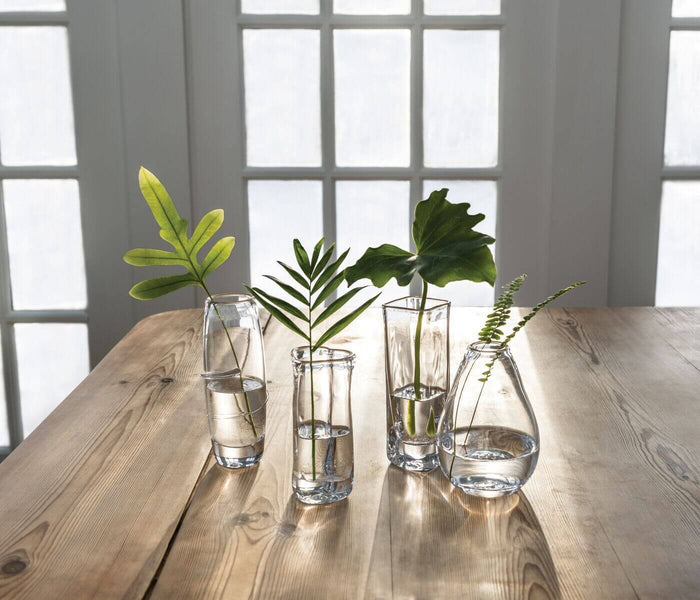 Four clear glass bud vases with water and fresh green foliage arranged on a wooden table near windows.
