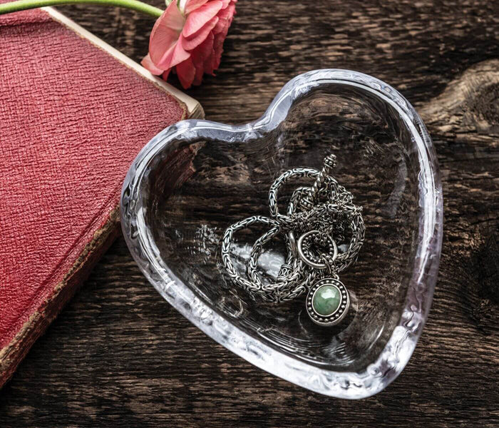Clear glass heart-shaped dish with ornate silver rings and green stone inside, on wooden surface.