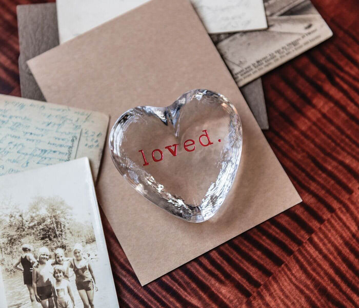 Clear glass heart-shaped object with "loved" text etched in red, displayed on tan paper.
