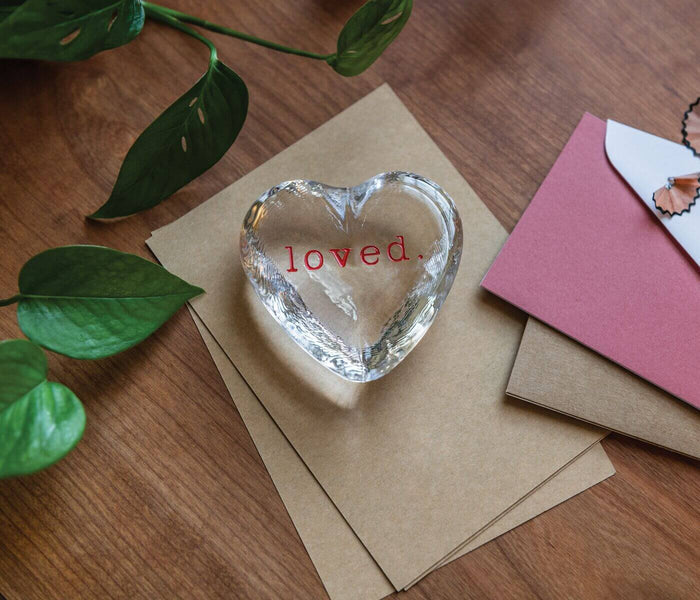 Clear glass heart-shaped object with "loved" text engraved on kraft paper, surrounded by plant leaves and greeting cards.