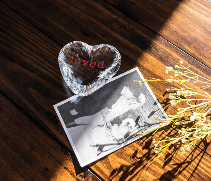 Clear glass heart with "loved" engraved in red text, placed on a black and white photograph atop a wooden surface.