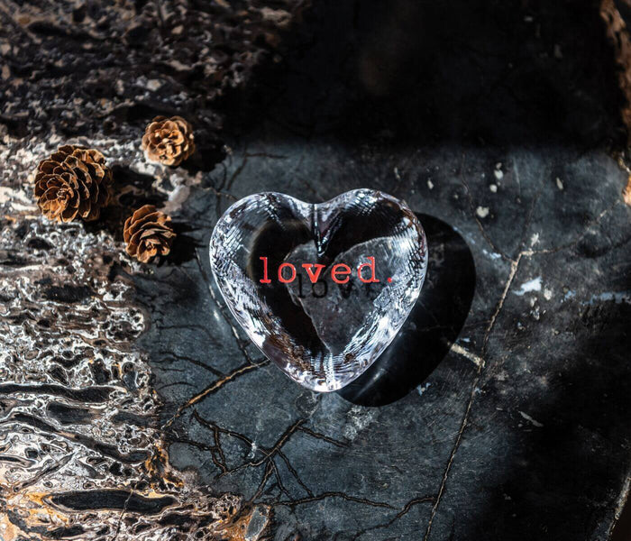 Clear glass heart with "loved" text, surrounded by dried pinecones and twigs on dark textured surface.