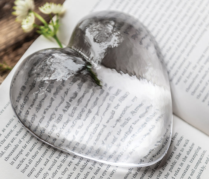 Clear glass heart-shaped paperweight with printed book text visible inside, displayed on open book pages with small white flo