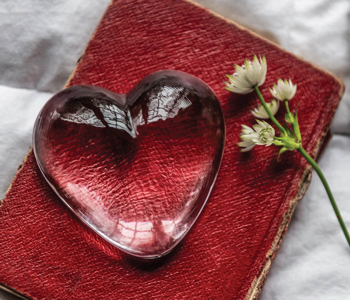 Clear glass heart-shaped paperweight on red textile with small white flowers beside it.