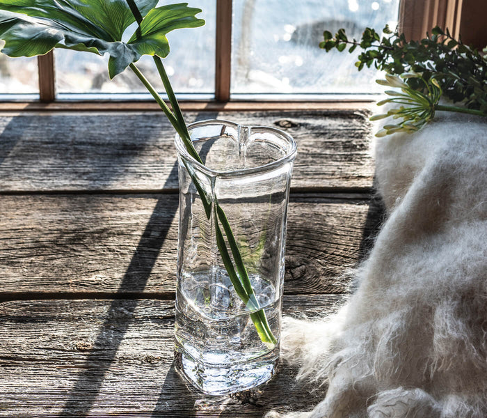 Clear glass vase with heart-shaped top holding a green stem, displayed on wooden surface by window.