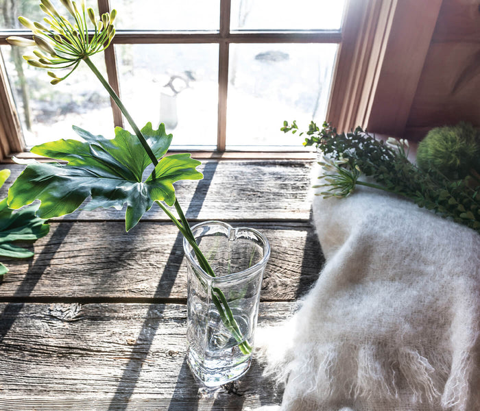 Clear glass vase with heart-shaped outline holding a single green stem on a wooden surface.