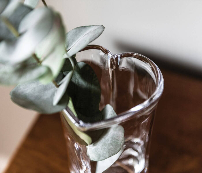 Clear glass heart-shaped vase with eucalyptus stems and gray-green leaves inside.