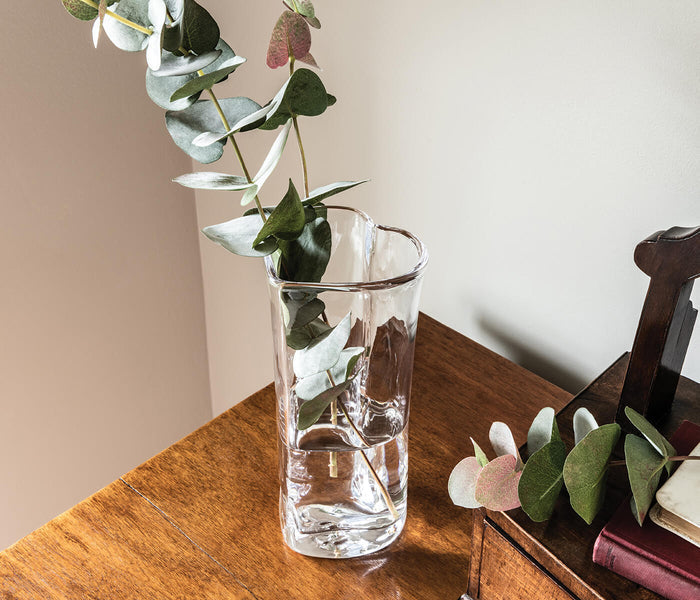 Clear glass vase with heart-shaped indentations holding eucalyptus stems and leaves on wooden surface.