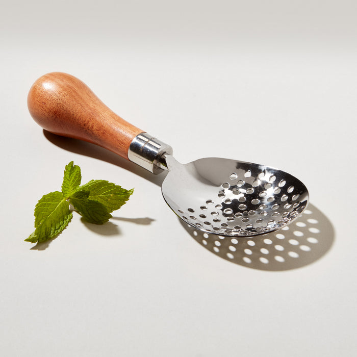 Silver-toned julep strainer with perforated bowl, wooden handle, and metal ferrule, displayed with fresh mint leaves.