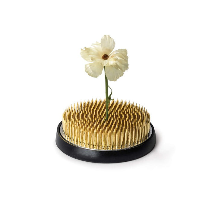 Brass kenzan flower frog with dense metal pins on black rubber base holding a white flower.