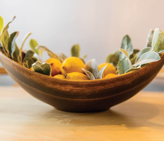 Dark walnut wooden bowl with live edge rim, filled with yellow fruits and green foliage.
