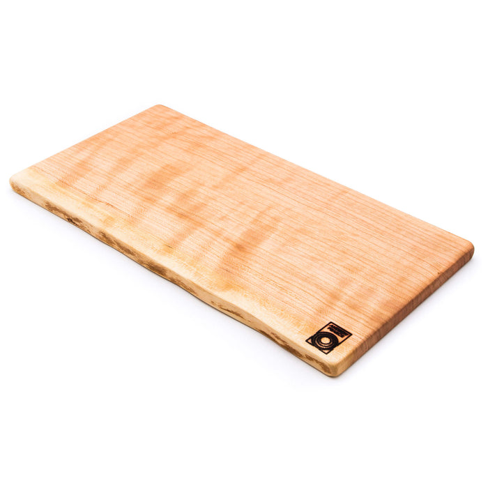 Cherry wood board with live edge and natural grain, featuring a small metal maker's mark on the lower right corner.